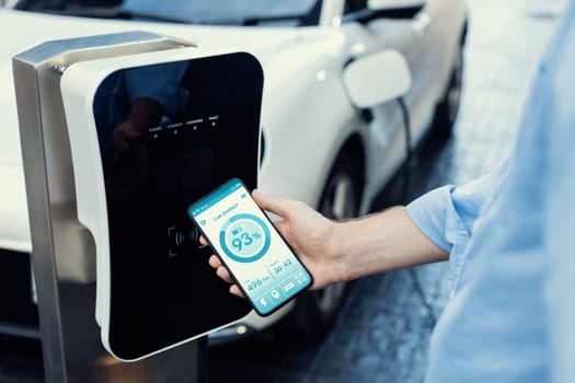 Closeup progressive businessman look at EV car's battery status application on smart phone screen at public parking charging station with power cable plug and renewable energy-powered electric vehicle