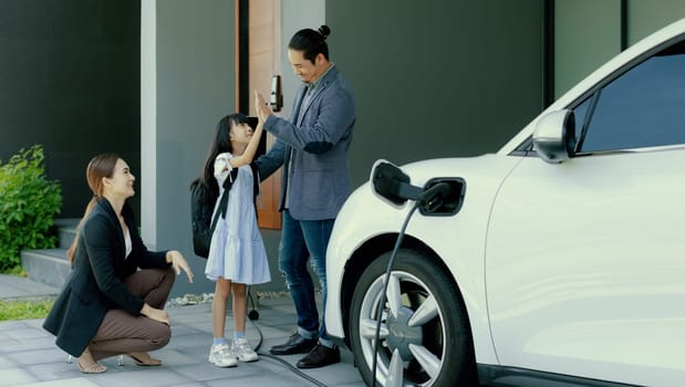 Progressive young parents and daughter with electric vehicle and home charging station. Green and clean energy from electric vehicles for healthy environment. Eco power from renewable source at home.