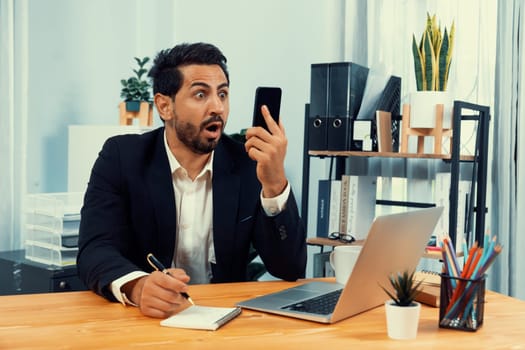 Diligent businessman busy talking on the phone call with clients while working with laptop in his office desk as concept of modern hardworking office worker lifestyle with mobile phone. Fervent