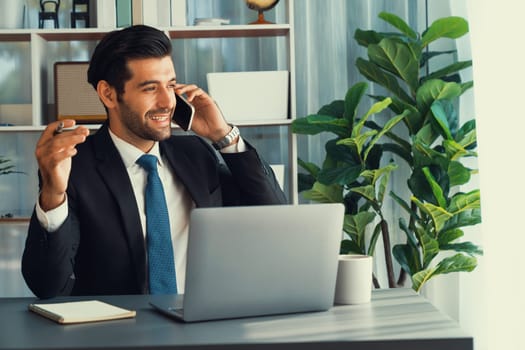Diligent businessman busy talking on the phone call with clients while working with laptop in his office as concept of modern hardworking office worker lifestyle with mobile phone. Fervent