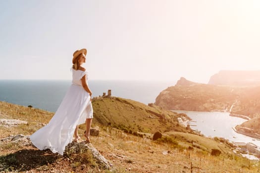 Happy woman in a white dress and hat stands on a rocky cliff above the sea, with the beautiful silhouette of hills in thick fog in the background