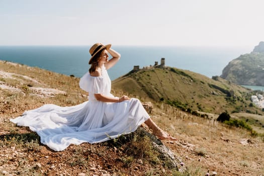 Happy woman in a white dress and hat stands on a rocky cliff above the sea, with the beautiful silhouette of hills in thick fog in the background