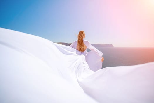 A blonde woman with long hair stands on a sunny seashore in a flowing white dress, with the silk fabric waving in the wind, and blue sky and mountains in the background
