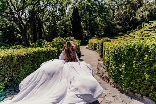 Brunette runs white dress park. A beautiful woman with long brown hair and a long white dress runs along the path along the beautiful bushes in the park, rear view.