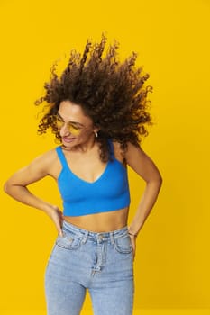 Woman with curly afro hair in a blue T-shirt on a yellow background dancing flying hair with sunglasses yellow, hand signs, look into the camera, smile with teeth and happiness, copy space. High quality photo