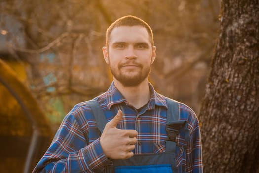 Farmer satisfied european appearance rural portrait at sunset with beard, shirt and overalls looking at camera, showing cool gesture with hand finger outdoors.