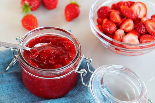 Glass jar with strawberry jam prepared for canning and fresh strawberries in a bowl on the table.