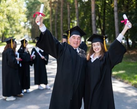 A group of graduates in robes outdoors. An elderly man and a young woman congratulate each other on their graduation