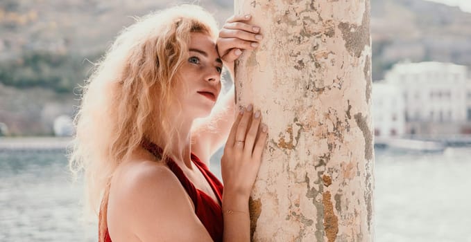 Close up shot of beautiful young caucasian woman with curly blond hair and freckles looking at camera and smiling. Cute woman portrait in a pink long dress posing on a volcanic rock high above the sea