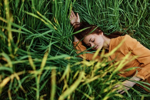 a close horizontal photo of a pleasant woman in a long orange dress resting lying in the tall grass with her eyes closed in sunny weather at sunset with her arms outstretched. Street photography, the theme of privacy with nature. High quality photo