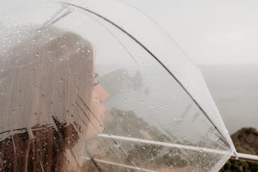 Woman rain park. Happy woman portrait wearing a raincoat with transparent umbrella outdoors on rainy day in park near sea. Girl on the nature on rainy overcast day