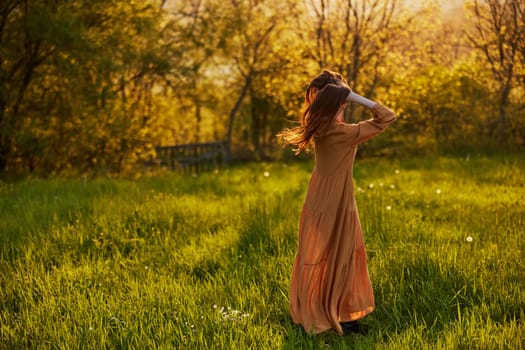 a slender woman with long hair stands in a field with her back to the camera, illuminated by the rays of the setting sun and happily poses enjoying the warm weather and rest. High quality photo