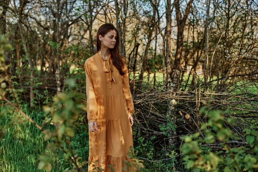 a beautiful, slender woman with long hair walks in the shade near the trees, dressed in a long orange dress, enjoying the weather and the weekend. The theme of privacy with nature, horizontal photography on the street. High quality photo