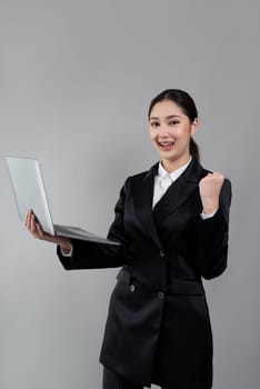 Confident young businesswoman stands on isolated background, working on laptop posing in formal black suit. Office lady or manager with smart and professional appearance. Enthusiastic