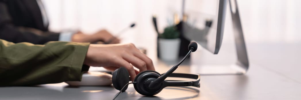 Panorama focus hand holding headset on call center workspace desk with blur background of operator team or telesales representative engaging in providing client with customer support service. Prodigy