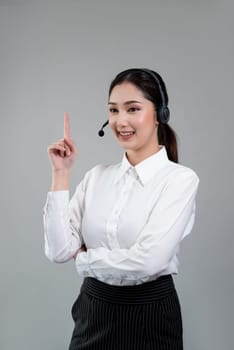 Asian female call center operator with smile face advertises job opportunity, wearing a formal suit and headset pointing finger for product on customizable isolated background. Enthusiastic