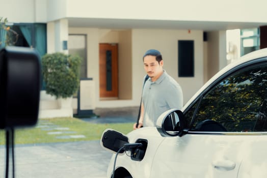 Progressive asian man install cable plug to his electric car with home charging station in the backyard. Concept use of electric vehicles in a progressive lifestyle contributes to clean environment.