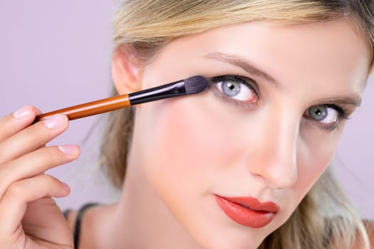 Closeup beautiful girl with flawless applying alluring eye shadow makeup with eyeliner brush. Cosmetic facial painting process on lovely young woman with perfect clean skin on pink isolated background