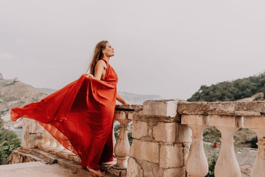 Side view a Young beautiful sensual woman in a red long dress posing on a volcanic rock high above the sea during sunset. Girl on the nature on overcast sky background. Fashion photo