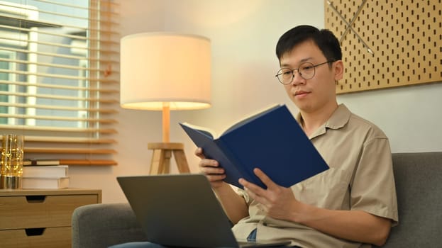 Peaceful asian man wearing glasses sitting on sofa and reading book. Leisure and people concept.