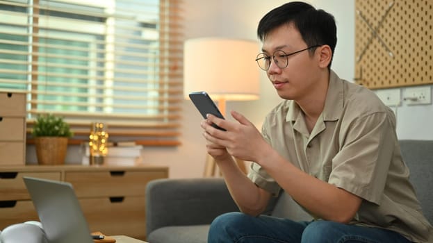 Image of adult asian man in casual watching video or playing game on smart phone, sitting on couch at home.