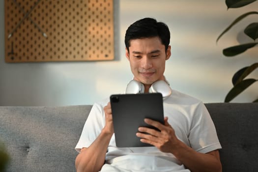 Handsome Asian man sitting on couch working remotely online on digital tablet at home.
