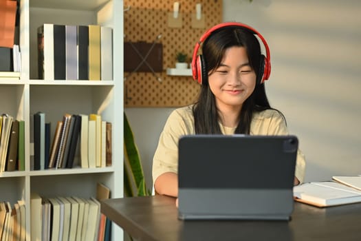 Smiling asian girl wearing headphones sitting at table learning online at virtual class on laptop. Distance education concept.