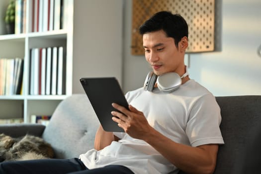 Millennial Asian man in casual clothes sitting on couch and using on digital tablet. People, technology and lifestyle.