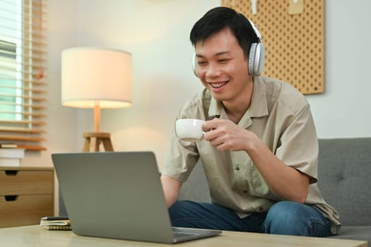 Pleasant man in wireless headphone and casual clothes drinking coffee and using laptop on couch at home.