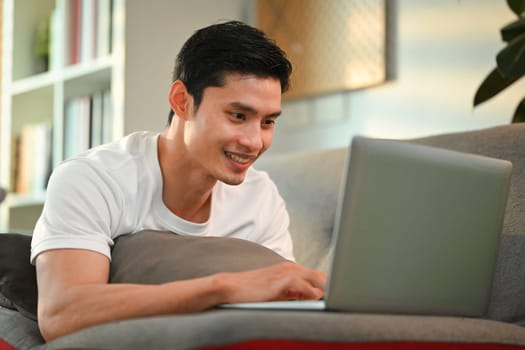 Happy adult asian man lying on couch and enjoy working on internet, chatting in social media on laptop.