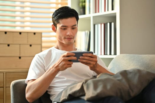 Happy millennial man playing video games on smart phone while resting on couch.Technology, entertainment and leisure activity.