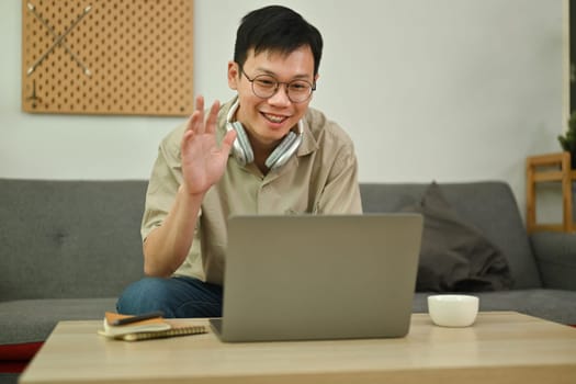 Handsome Asian man making video call on laptop sitting on couch at home. Technology, communicating and lifestyle.