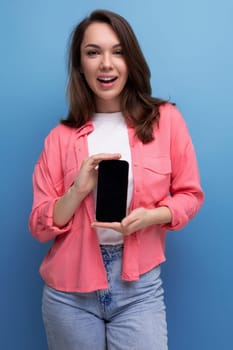 a smiling brunette lady with dark hair below her shoulders in a shirt and jeans demonstrates a smartphone screen forward.