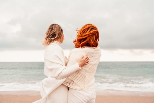 Women sea walk friendship spring. Two girlfriends, redhead and blonde, middle-aged walk along the sandy beach of the sea, dressed in white clothes. Against the backdrop of a cloudy sky and the winter sea. Weekend concept