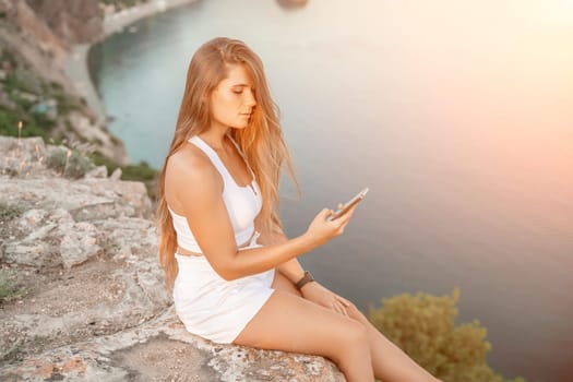 Happy woman in white shorts and T-shirt, with long hair, talking on the phone while enjoying the scenic view of the sea in the background