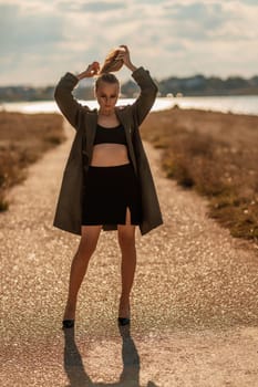 Portrait blonde sea cape. A calm young blonde in an unbuttoned khaki raincoat walks along the seashore, under a raincoat a black skirt and top.