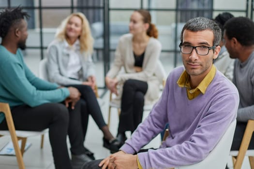 A happy young businessman, in a purple sweater, sits against the background of his colleagues in a modern office