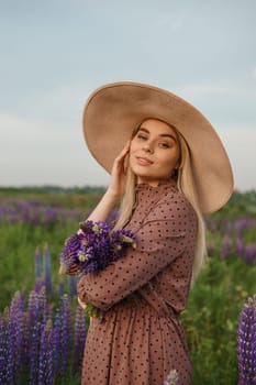 A beautiful woman in a straw hat walks in a field with purple flowers. A walk in nature in the lupin field.