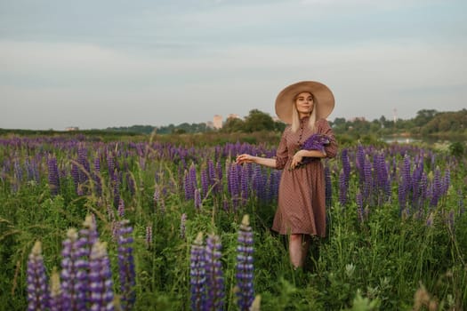 A beautiful woman in a straw hat walks in a field with purple flowers. A walk in nature in the lupin field.