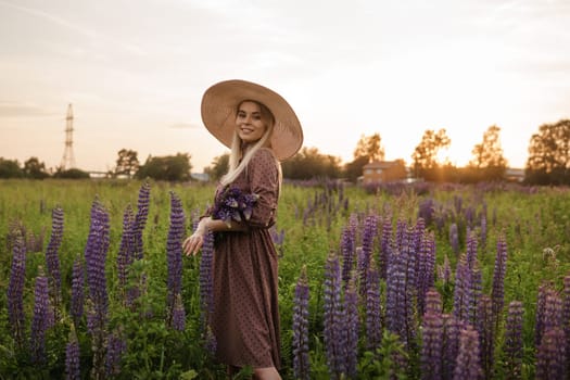 A beautiful woman in a straw hat walks in a field with purple flowers. A walk in nature in the lupin field.