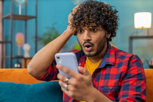 Sad displeased indian man use smartphone typing browsing, loses becoming surprised sudden lottery results, bad news, fortune loss, fail, deadline, virus. Young hindu guy at home living room on couch