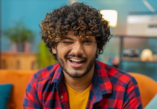 Smiling playful happy young indian man blinking eye, looking at camera with smile, winking and flirting, expressing optimism. Portrait of hindu guy at home apartment living room sitting on couch