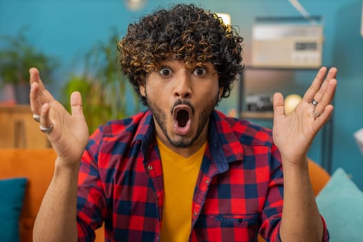 Oh my God, Wow. Young indian man surprised looking at camera with big eyes, shocked by sudden victory, good win news, celebrating. Portrait of excited amazed hindu guy at home apartment room on sofa
