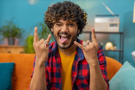 Overjoyed delighted young indian man showing rock n roll gesture by hands, cool sign, shouting yeah with crazy expression, dancing. Hindu guy emotionally rejoicing in success at home living room couch