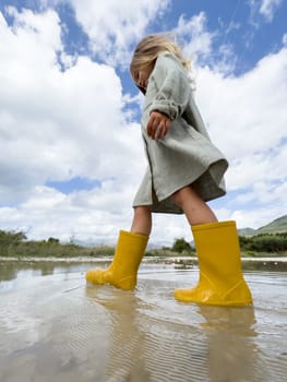 Little girl in yellow rubber boots walks through a puddle against a cloudy sky. High quality photo