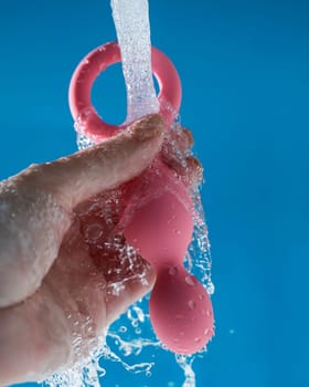 Woman holding pink anal beads under running water on blue background. Sex toy hygiene concept