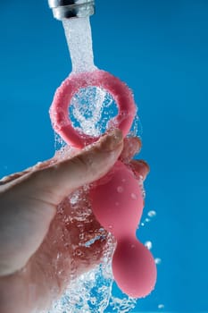 Woman holding pink anal beads under running water on blue background. Sex toy hygiene concept