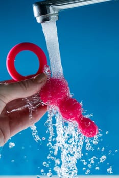 Woman holding pink anal beads under running water on blue background. Sex toy hygiene concept