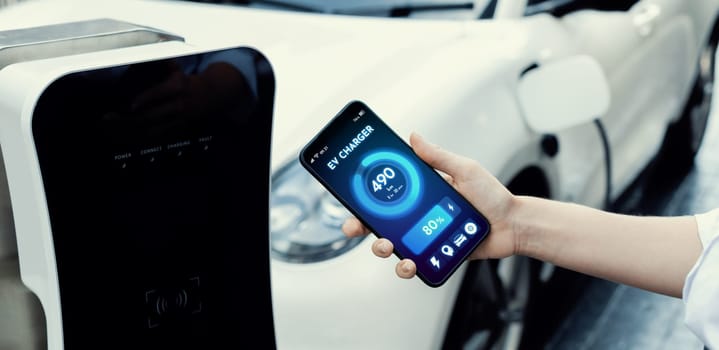 Battery status of electric vehicle displayed on smartphone application or software while vehicle is plugged into charging station for progressive future refueling. Battery status on phone screen.