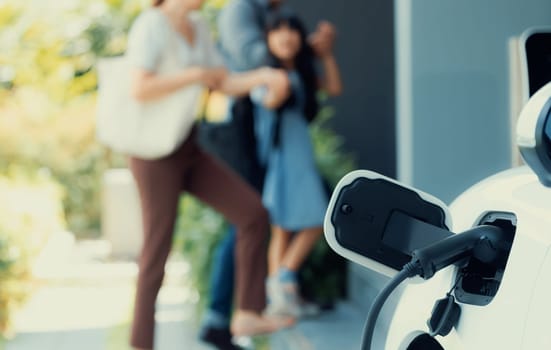 Focus closeup electric vehicle recharging battery from home electric charging station with blurred family in background. Renewable clean energy car for progressive eco awareness lifestyle concept.